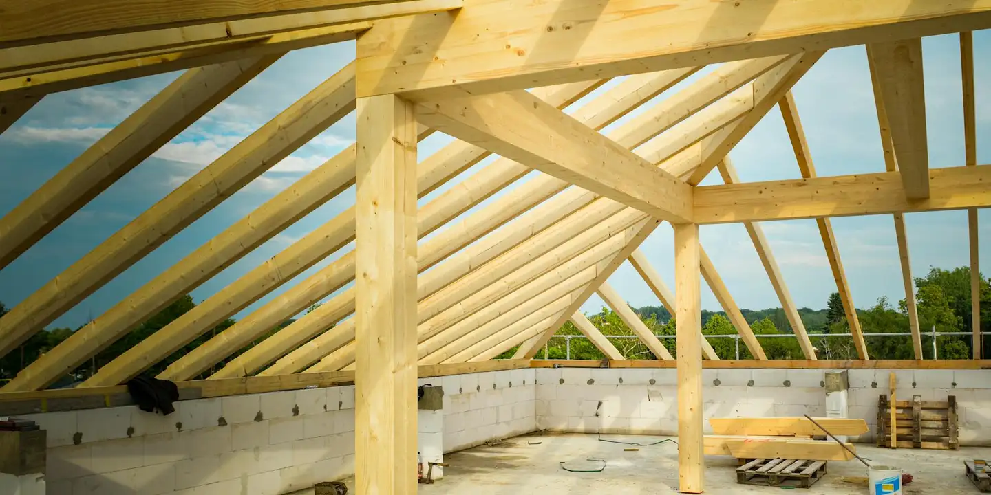 Interior view of a wooden roof frame with rafters and a central post, daylight streaming through the framing.