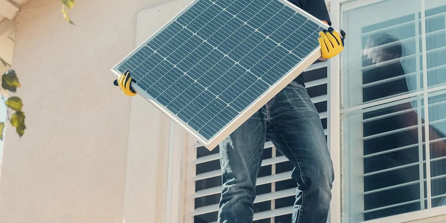 A worker wearing yellow gloves holds a solar panel near a window, illustrating rooftop work.