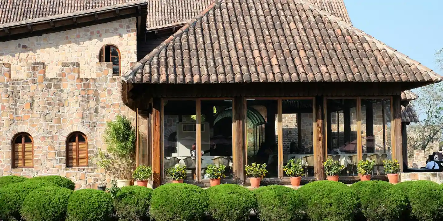 Stone house with a clay-tiled roof and a glass-enclosed sunroom, bordered by a row of potted plants.