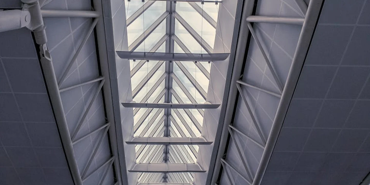 View of exposed roof trusses with a skylight, showing a high, complex framework and daylight streaming through the structure.