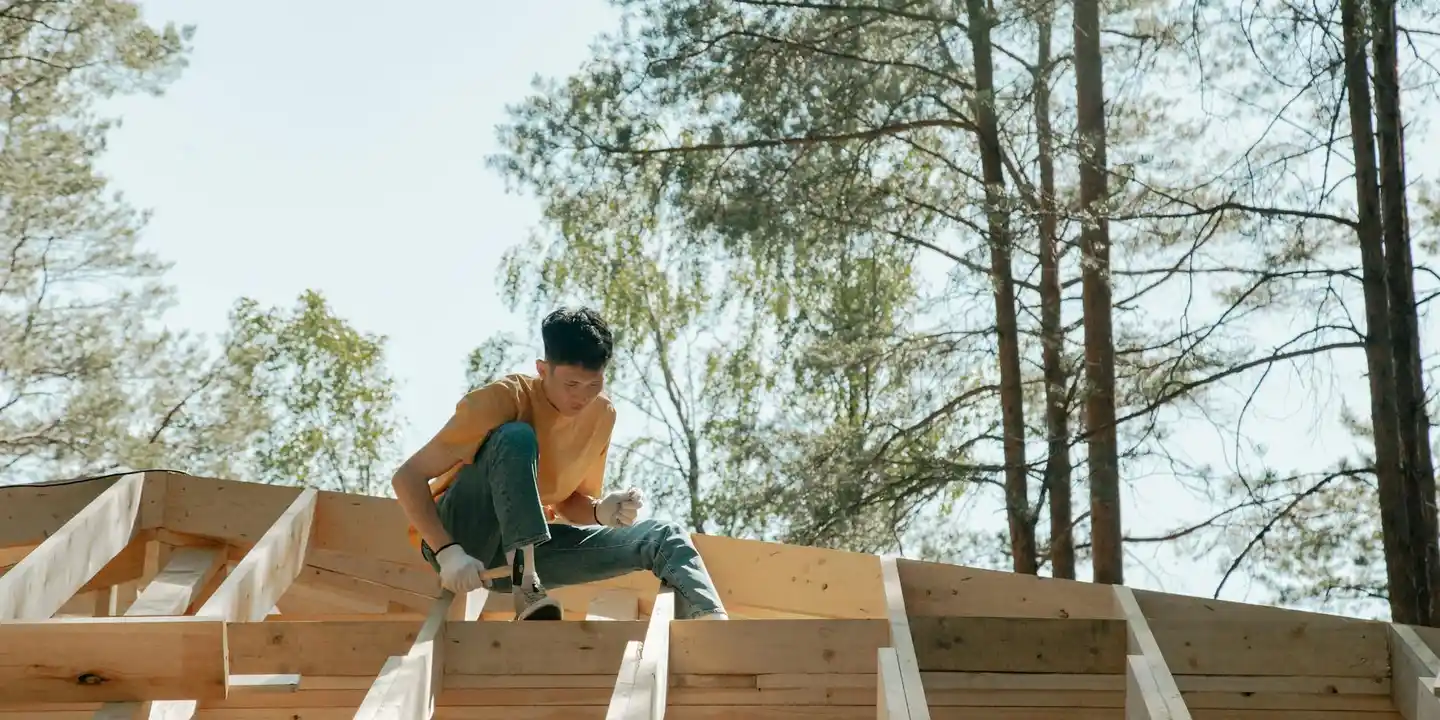 Construction worker perched on a wooden residential roof deck frame, preparing to install roofing felt underlayment in a sunny, wooded area.