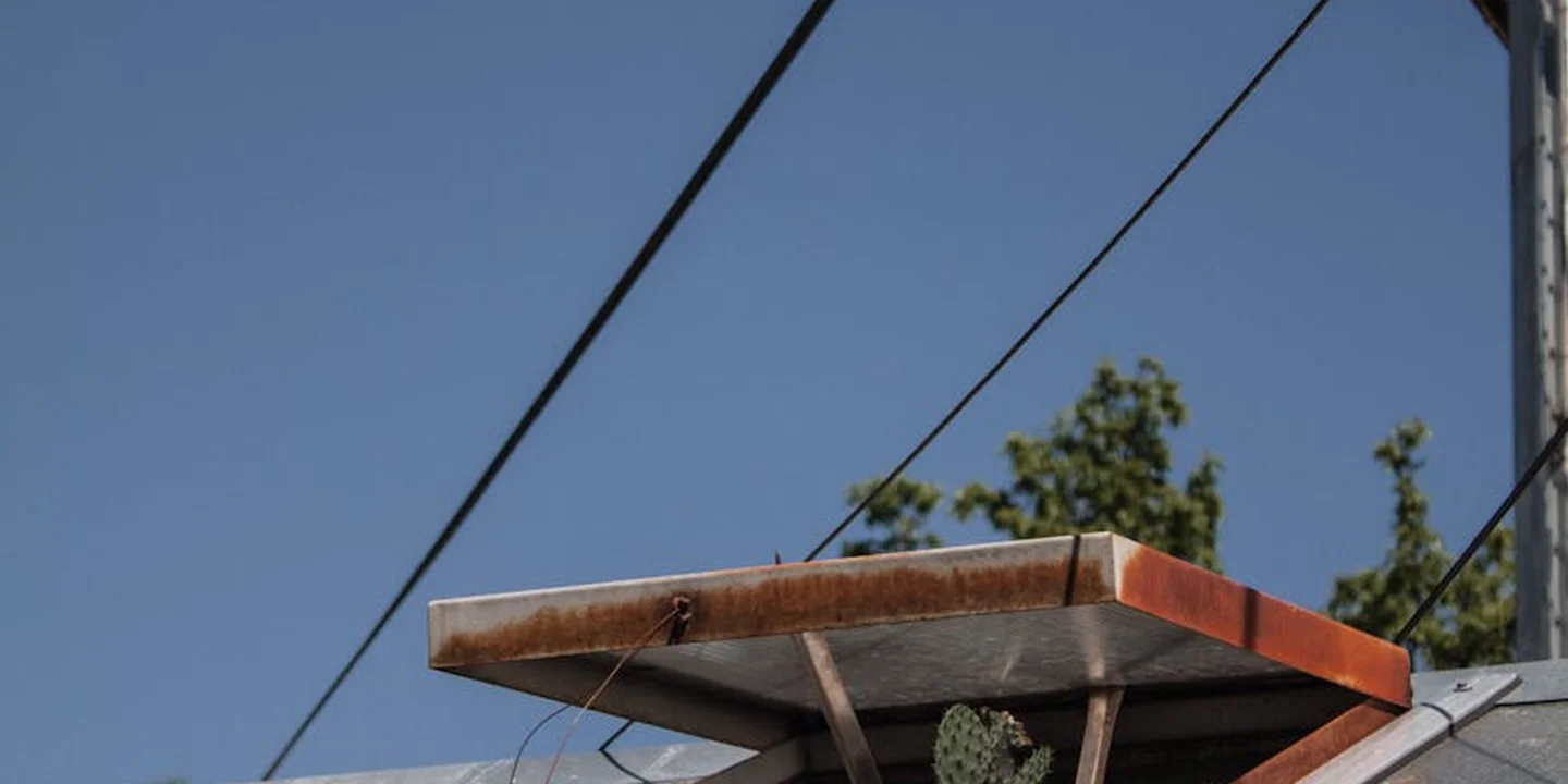 Rusty metal roof vent protruding from a sloped roof with a clear blue sky and overhead power lines in the background