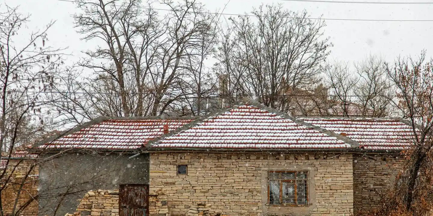 A small stone house with a snow-covered roof and bare trees in the background.