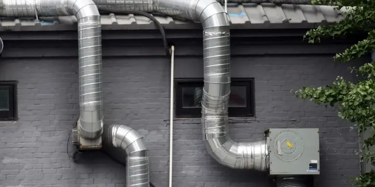 Exterior wall with shiny silver metal ventilation ducts and a small electrical unit, illustrating roof/attic ventilation components.
