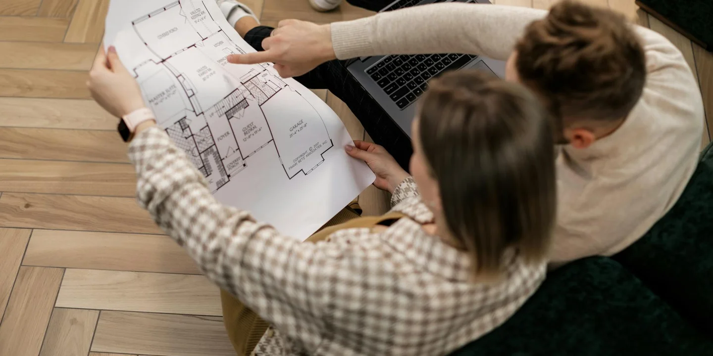 Group of people studying a roof blueprint spread on a wooden floor, with a laptop nearby