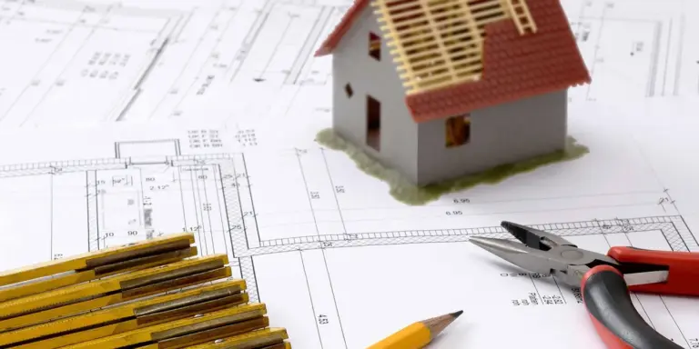 A small model house sits on top of architectural roof blueprints with drafting pencils and pliers nearby.