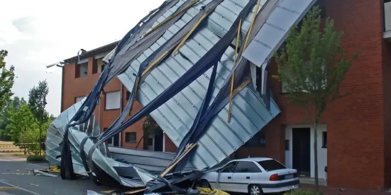 Severely damaged metal roofing panels collapsed over a red-brick building, with a parked sedan and trees in the background.