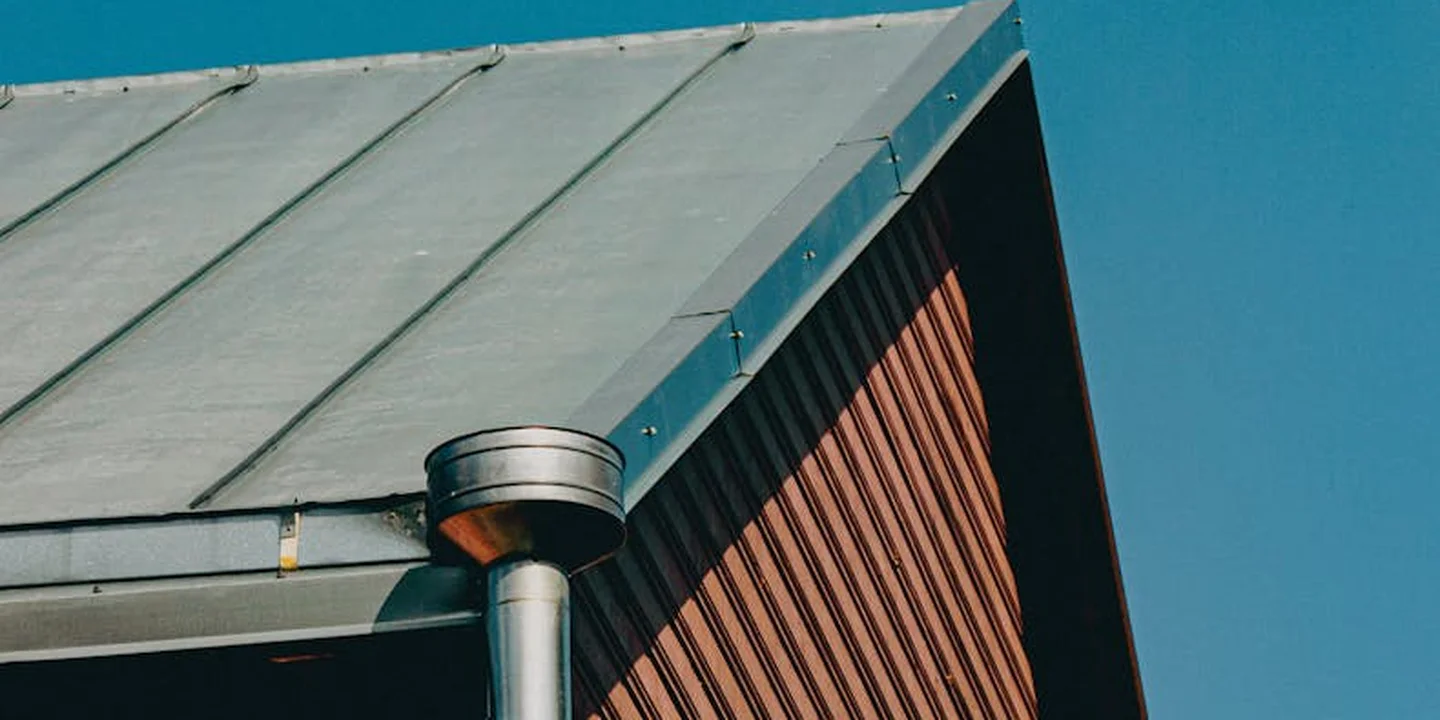 Close-up of a metal roof edge with a gutter and a circular vent near the roof, set against a clear blue sky.