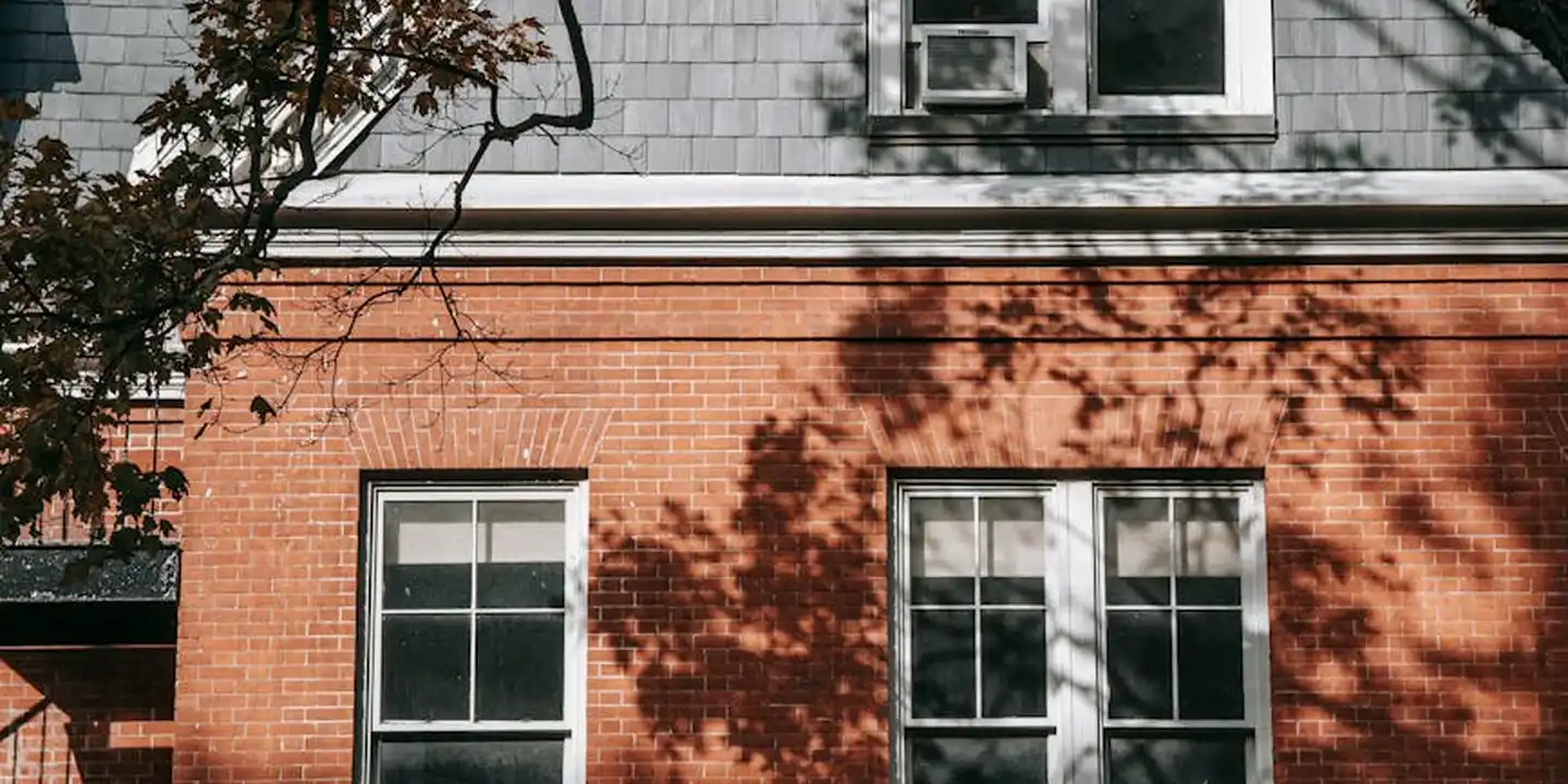 A brick residence with two white-framed windows on the lower level, tree shadows across the wall, and a roof edge with white trim visible above, illustrating roof-to-wall junctions where leaks commonly begin.