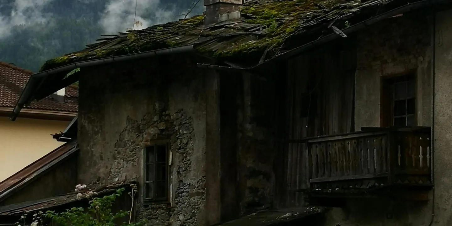 Weathered residential roof with patches of green moss and dark, aging shingles, with an old exterior wall nearby.