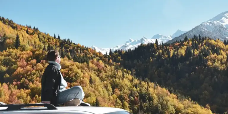 Person sitting on top of a car's roof rack overlooking colorful autumn trees and snow-capped mountains