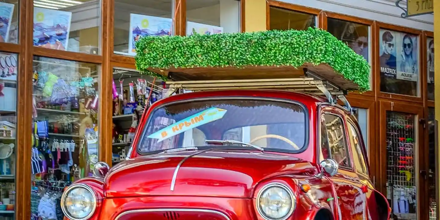 A red vintage car with a roof rack carrying a green cargo box parked in front of a storefront.