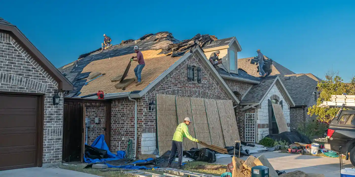 Roofing crew on a brick house repairing the roof; plywood underlayment, shingles and tools visible against a bright blue sky.