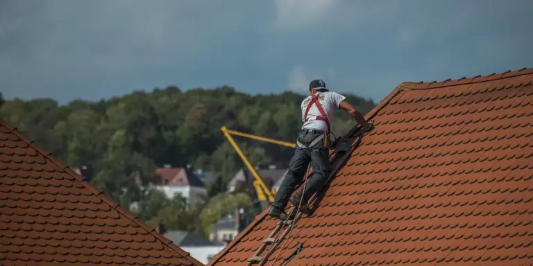 Construction worker in a safety harness repairing a tiled sloped roof with a crane in the background