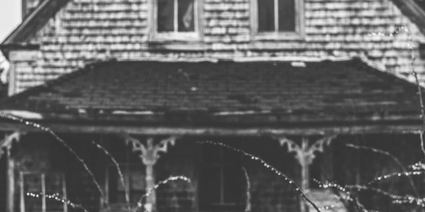 Black-and-white photograph of a two-story house with a pitched roof and a decorative front porch, suggesting elevated roofing work.
