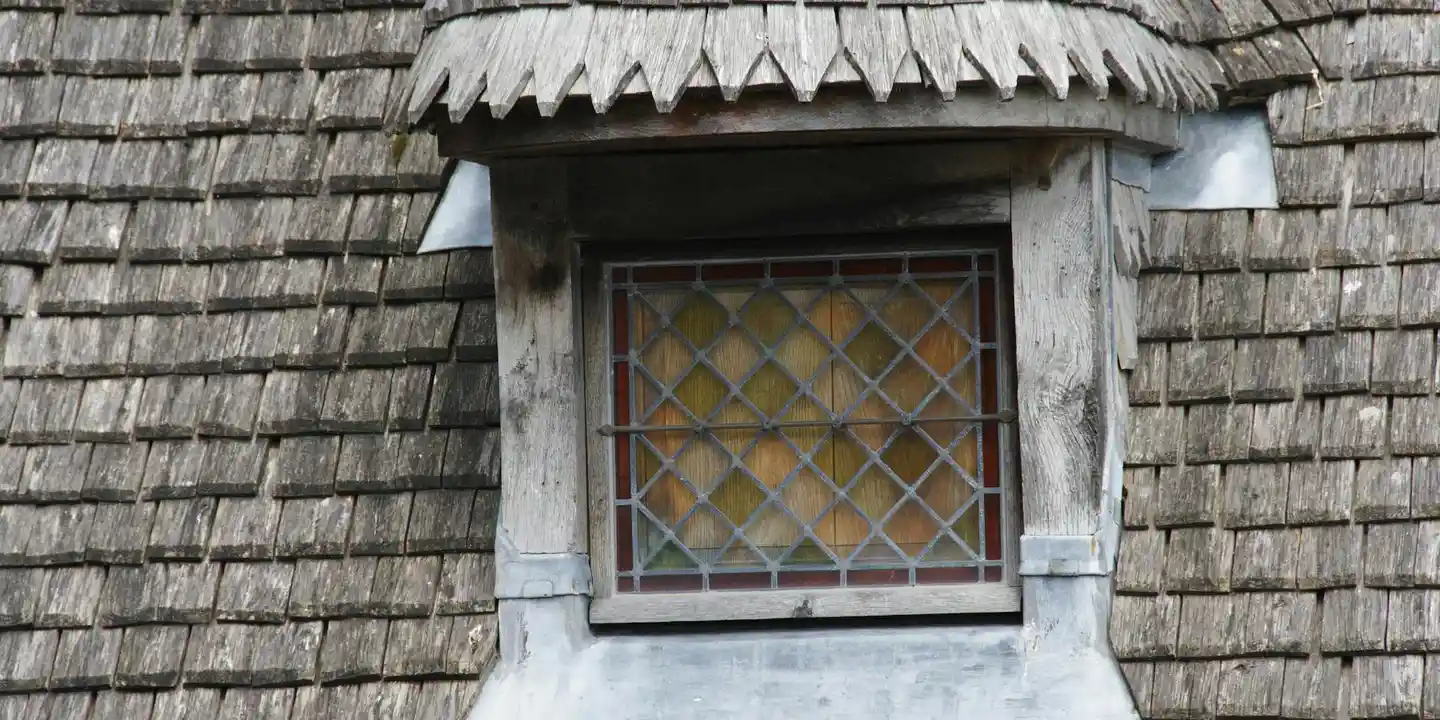 Close-up of weathered wooden roof shingles around a dormer window with lattice panes.