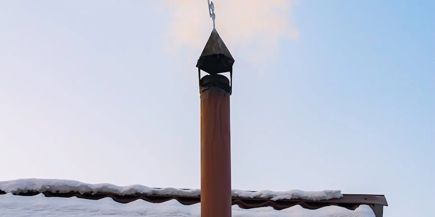 Snow-covered roof edge with a vertical vent pipe protruding from the roof under a pale blue sky.