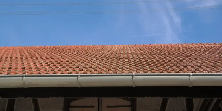 A house roof edge with a white gutter and gutter guard installed under red clay tile roofing, against a clear blue sky.