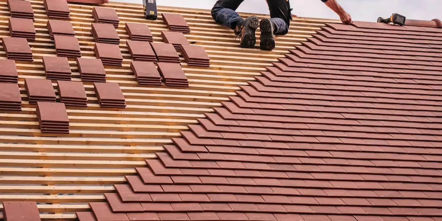 Two workers on a pitched roof installing brown shingles over wooden battents, with sections of shingles stacked on the left and visible flashing along the roof edge.