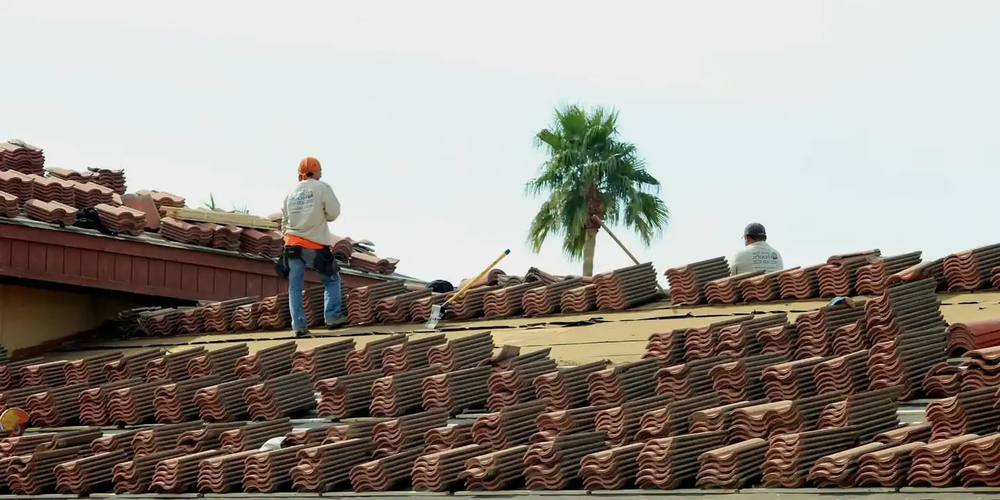 Two workers on a sloped roof installing reddish-brown tiles, with a palm tree in the background.