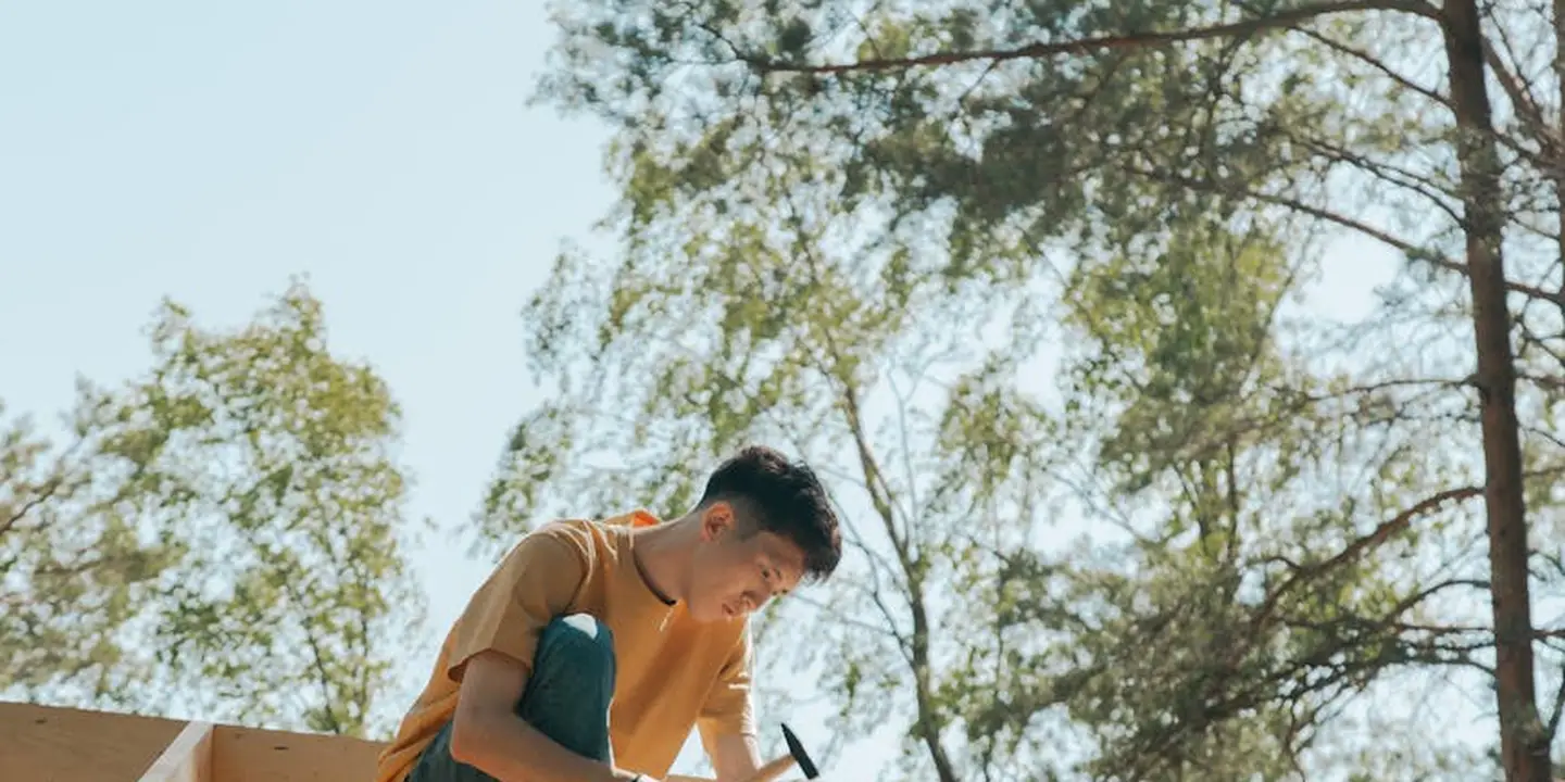 Person on a roof using a hammer, with trees and clear sky in the background
