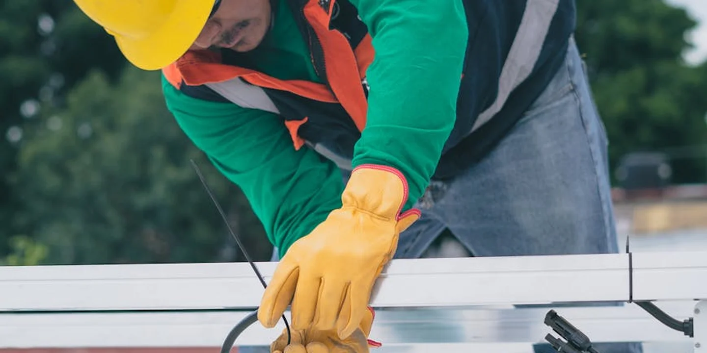 Construction worker wearing a yellow hard hat and orange gloves inspecting roofing materials on a sunny site.