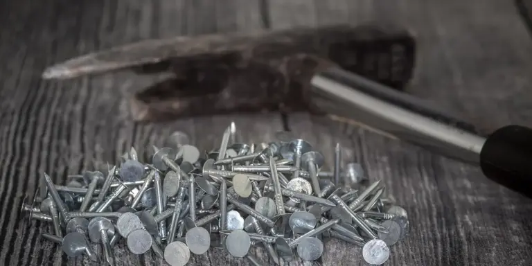 Close-up of a pile of metal roofing nails on a weathered wooden surface with a hammer in the background.