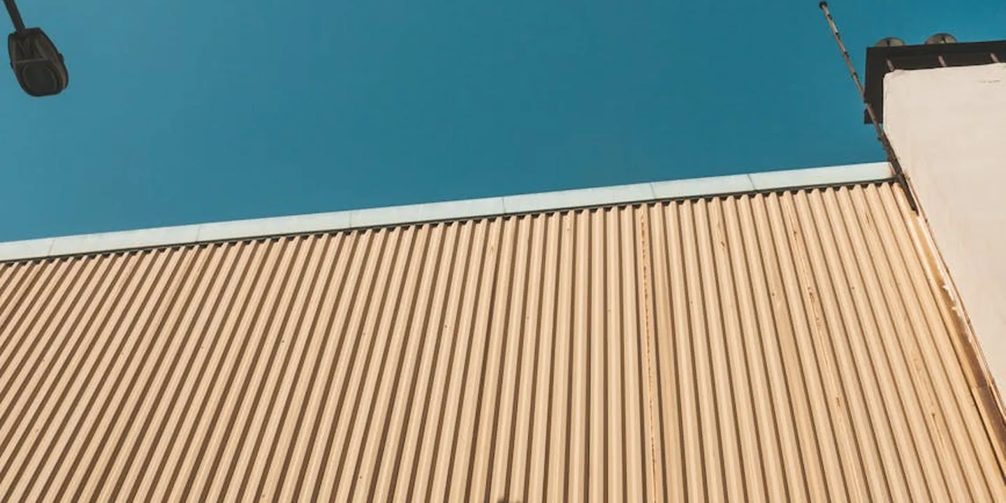 Side view of a pitched roof with tan metal panels against a clear blue sky, illustrating the final step of converting roof area from square feet to roofing squares.