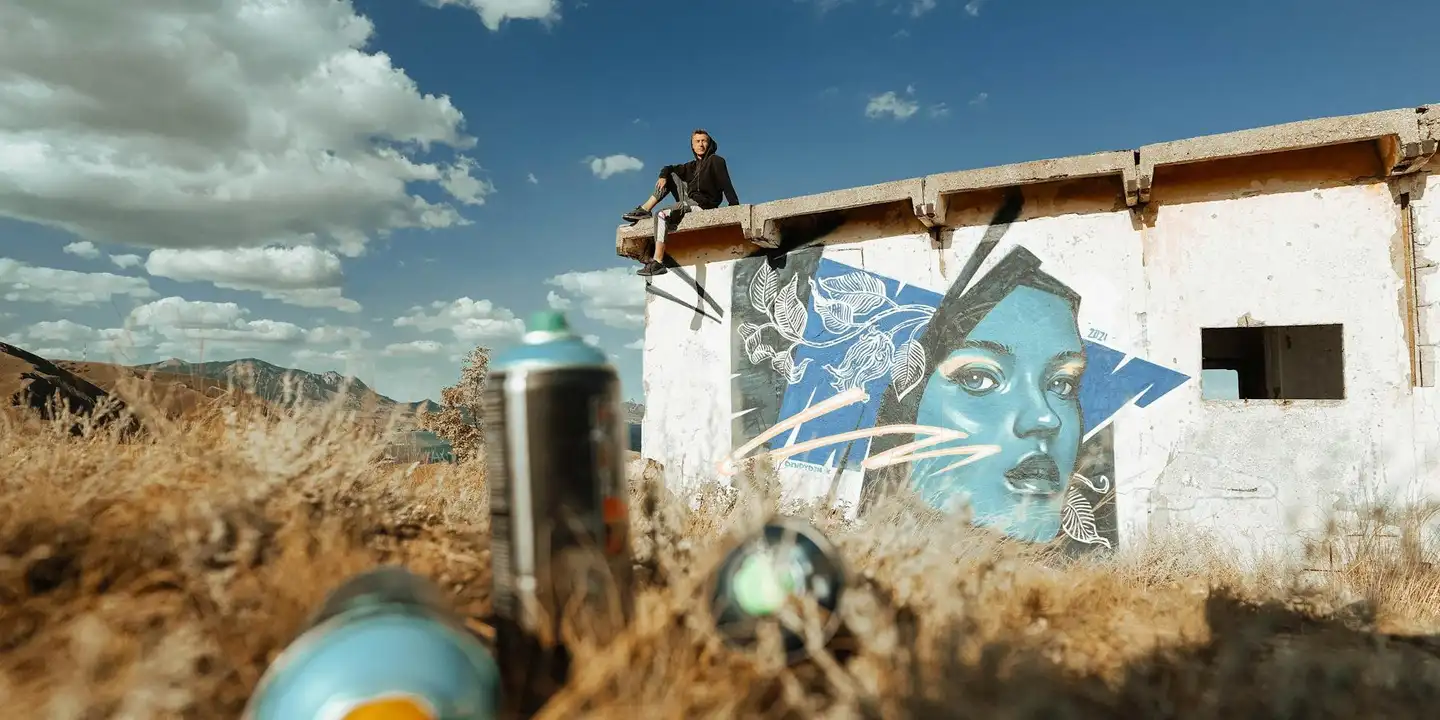 A person stands on a rooftop near a small white building with a blue graffiti mural; a spray paint can rests in the foreground.