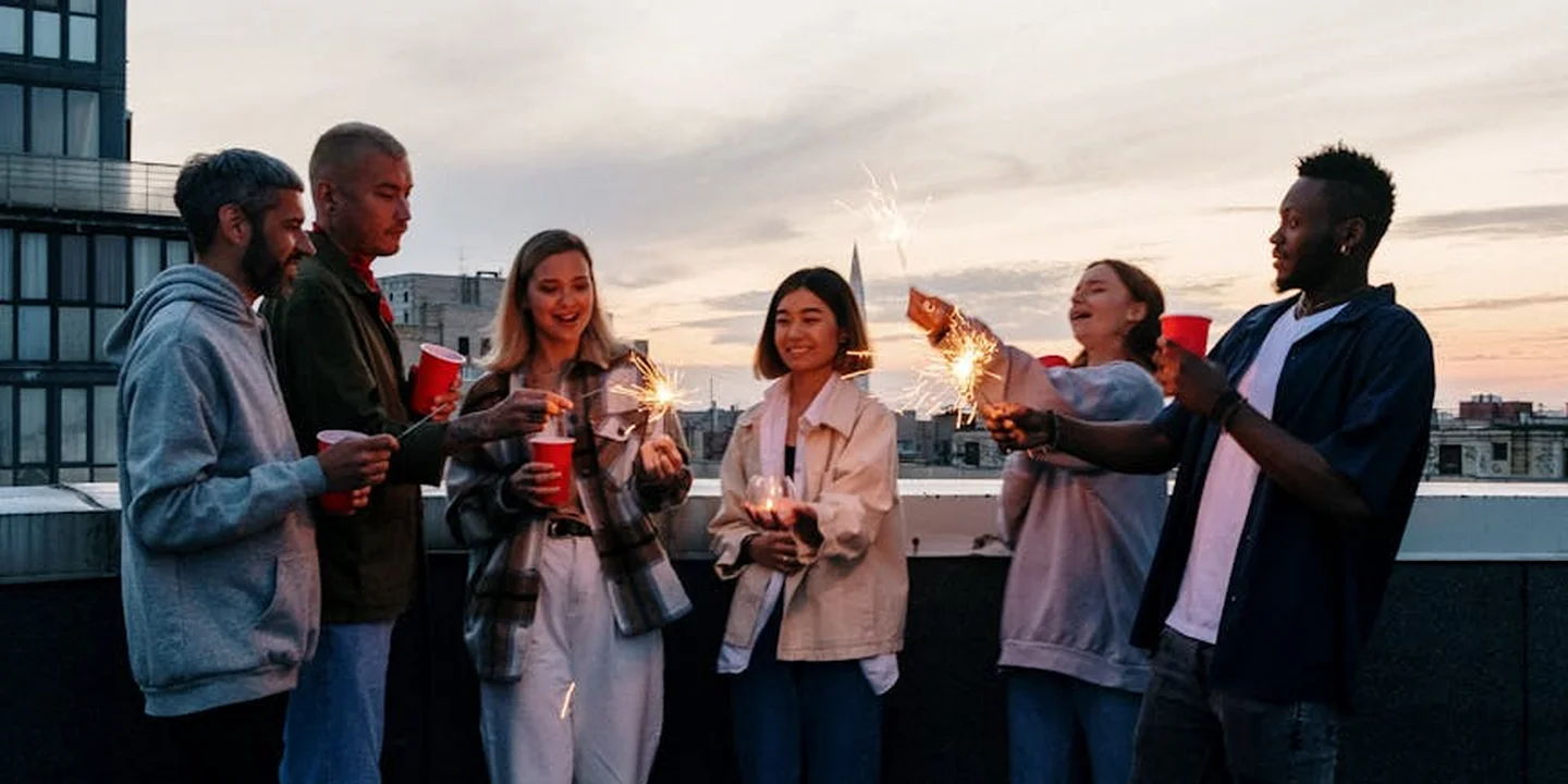 Diverse group of people on a rooftop at sunset, holding sparklers and drinks, engaging in conversation.
