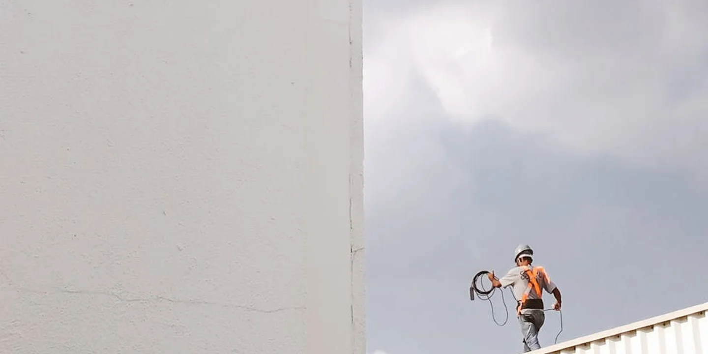 A roofer wearing a hard hat and orange shirt stands on a roof with a safety harness and rope against a cloudy sky.