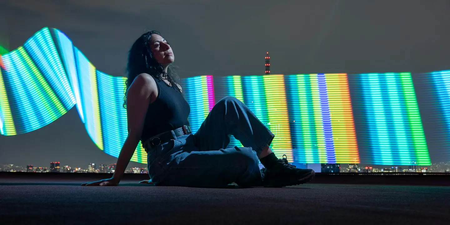 Person sitting on a rooftop at night with a multicolored LED light curtain in the background.