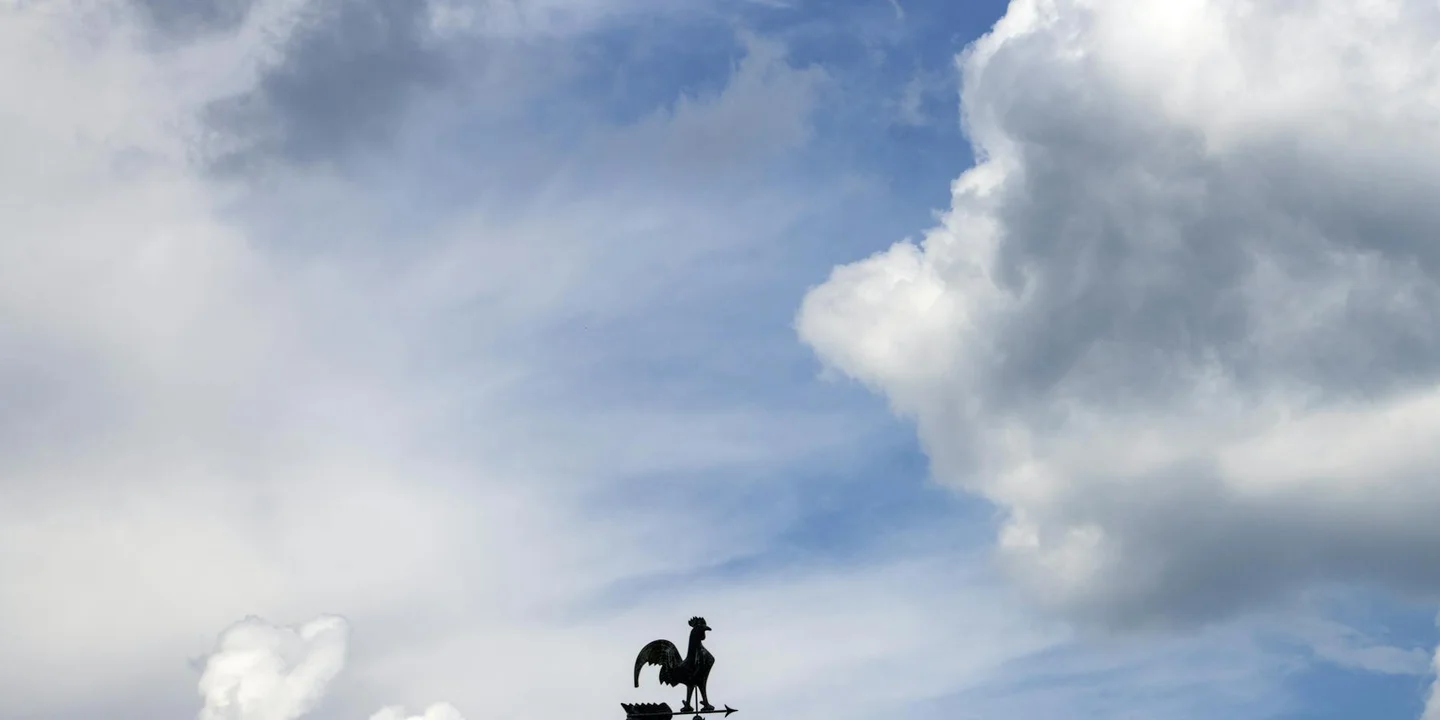Silhouette of a rooster weather vane on a residential roof against a blue sky with clouds.