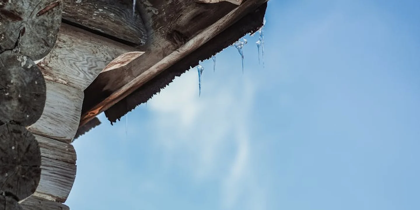 Close-up of a wooden roof edge with icicles against a clear blue sky, highlighting vulnerable gaps and flashing points that can cause leaks in rubber roofing.