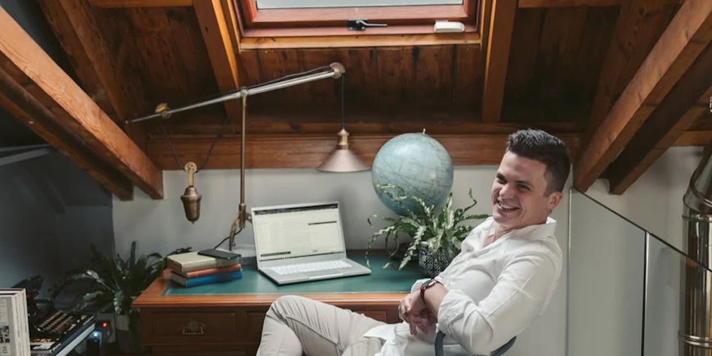 Attic workspace with a skylight, a man seated at a desk with a laptop, globe, and desk lamp preparing for skylight waterproofing work.