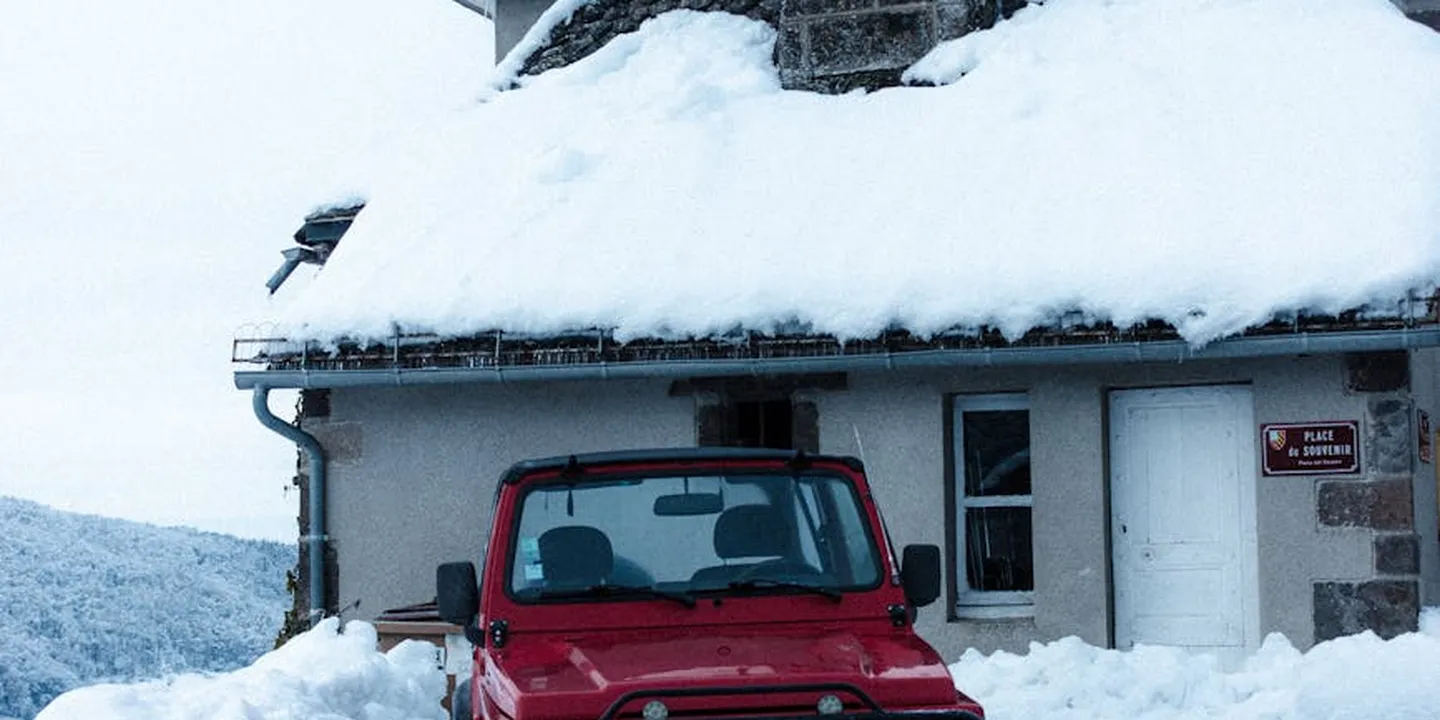 Red vehicle in front of a snow-covered house with a thick layer of snow on the roof and icicles along the eaves.