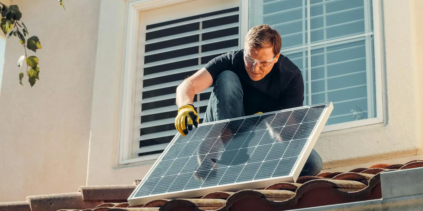 A worker wearing gloves mounts a solar panel on a residential tiled roof near a window.