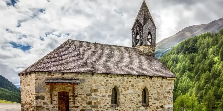 Rustic stone church with a bell tower set in a green mountain valley under a cloudy sky.