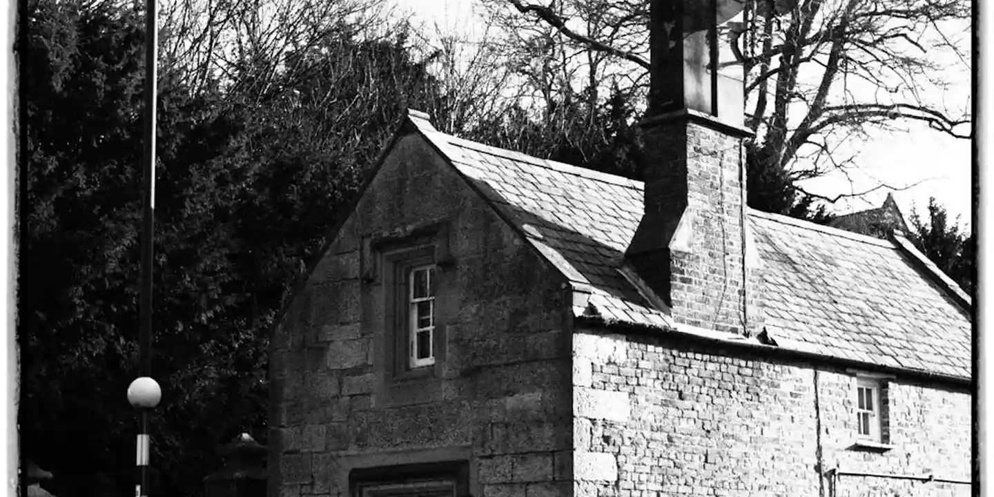 Black-and-white image of a stone cottage with a tall chimney and nearby trees.