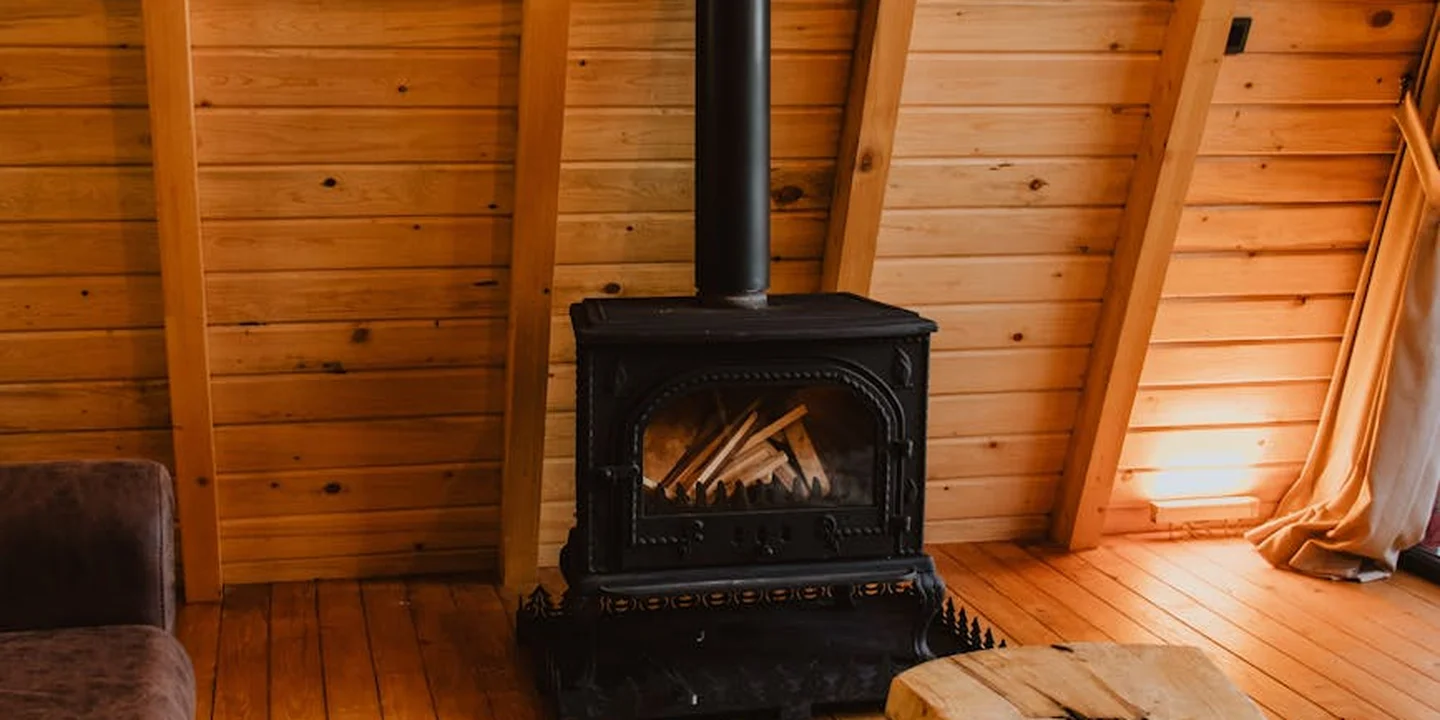 Wood stove in an attic with exposed wooden beams and sloped walls; a stovepipe rises from the stove toward the ceiling.
