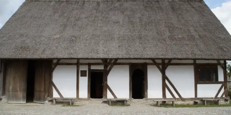 Traditional timber-framed cottage with a steep thatched roof and white plaster infill, with wooden benches in front.