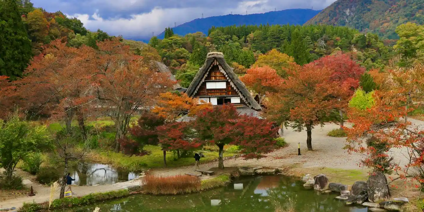 Autumn landscape with a traditional thatched-roof building among colorful trees and a tranquil pond.