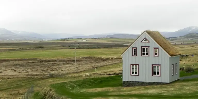 A small gray wooden house with a thatched roof sits in a wide open rural landscape of green fields, with distant mountains on the horizon.
