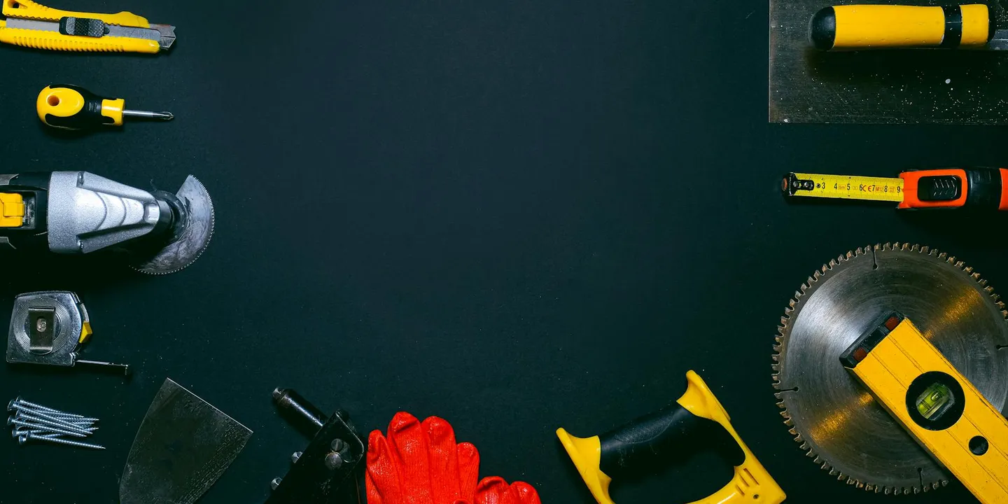 Top-down view of assorted roofing tools on a dark work surface, including a circular saw blade, tape measure, red work gloves, screw gun, bits, and a utility knife, prepared for sealing screws on a residential metal roof.