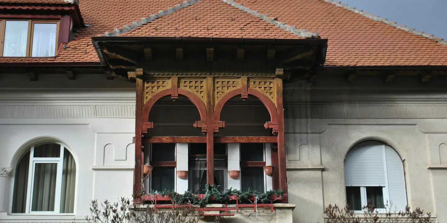White house with a decorative red wooden balcony featuring arches and planters beneath a red-tiled roof.