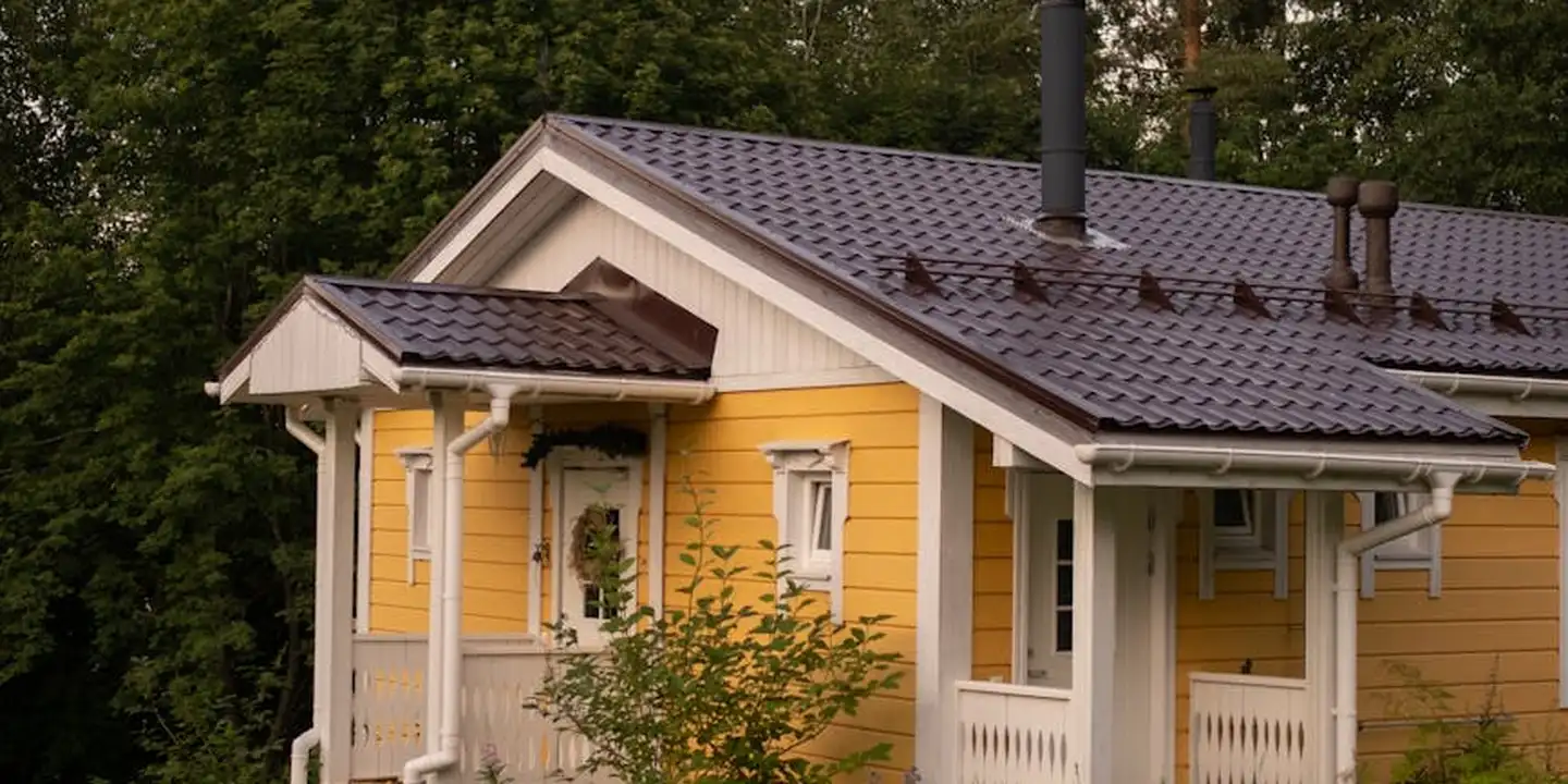 A yellow wooden house with a dark metal roof and a tall chimney, surrounded by trees.