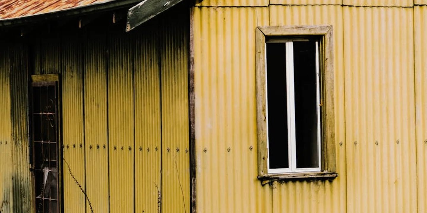 Weathered yellow corrugated metal siding with a small window, showing aging and wear on the roof and structure.