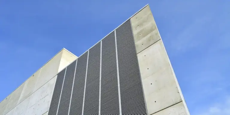 Diagonal view of a modern building facade with vertical perforated metal louvers, representing vent openings and airflow control.