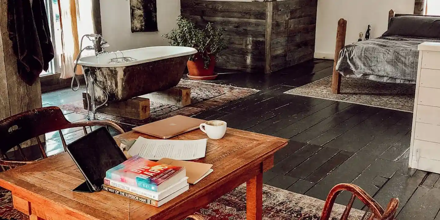 An attic workspace with a wooden desk, laptop, books, and papers; a bathtub and bed appear in the background, all on dark wooden flooring and exposed beams.