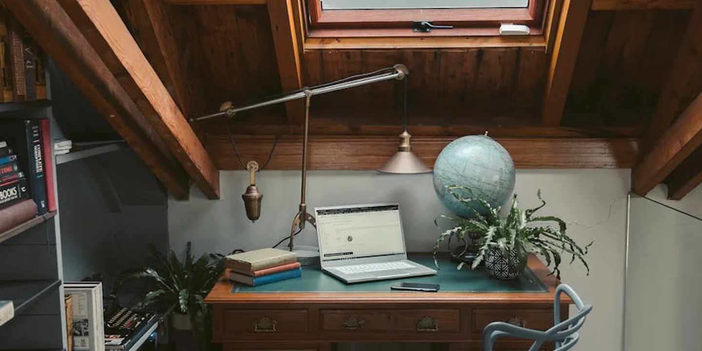Cozy attic workspace with a wooden desk, open laptop, globe, and potted plants beneath a skylight.